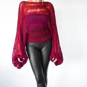 A burgundy open-knit sweater with long, voluminous sleeves. The sweater has a loose, oversized fit and is paired with black leather-look leggings. The top has a gradient effect, transitioning from a lighter red to a deeper burgundy.