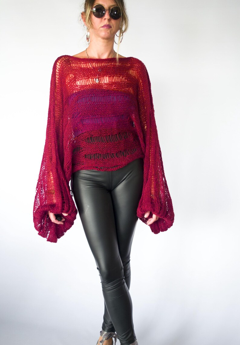 A burgundy open-knit sweater with long, voluminous sleeves. The sweater has a loose, oversized fit and is paired with black leather-look leggings. The top has a gradient effect, transitioning from a lighter red to a deeper burgundy.