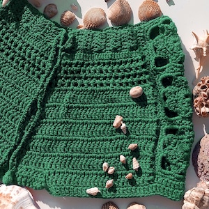 May include: A handmade, emerald green crochet piece with a textured pattern and scalloped edges. The crochet item is surrounded by various seashells and small stones, creating a beachy aesthetic. The piece is likely a shawl or a similar garment.