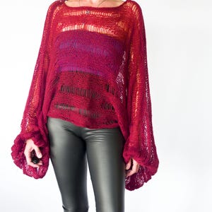 A loose-fitting, open-weave red sweater with long, flared sleeves. The sweater has a square neckline and a layered design, with a darker red and purple underlayer visible. The model is wearing black leggings.