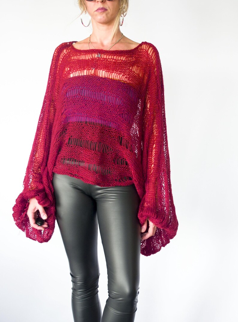 A loose-fitting, open-weave red sweater with long, flared sleeves. The sweater has a square neckline and a layered design, with a darker red and purple underlayer visible. The model is wearing black leggings.