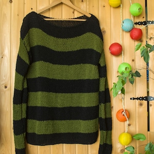 Op de afbeelding: Een gestreepte trui met zwarte en groene horizontale strepen. De trui hangt aan een houten hanger tegen een achtergrond van houten panelen.