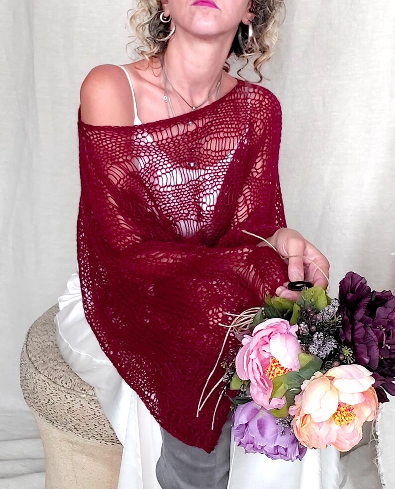 May include: A burgundy-colored, open-weave shawl draped over the shoulders. The shawl has a delicate, lacy pattern. A bouquet of pink, purple, and green flowers is held in the hand. The shawl is paired with a white top.
