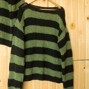 Green and black striped knit sweater hanging on wood panel background, showcasing its handcrafted design and punk-inspired vibe.