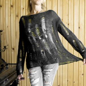 Black open-weave, heavily distressed knit sweater paired with ripped gray jeans; model with pink sunglasses and hoop earrings, wooden wall backdrop