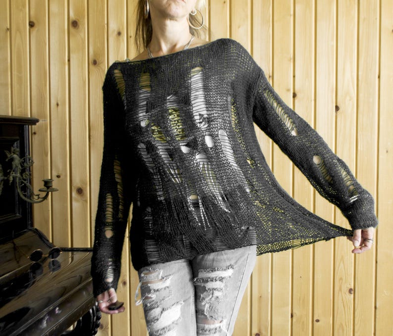 Black open-weave, heavily distressed knit sweater paired with ripped gray jeans; model with pink sunglasses and hoop earrings, wooden wall backdrop