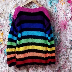 May include: A rainbow striped sweater with a pink neckline and black stripes. The sweater is hanging on a wooden hanger against a colorful floral background. The sweater's stripes include red, orange, yellow, green, blue, purple, and black.