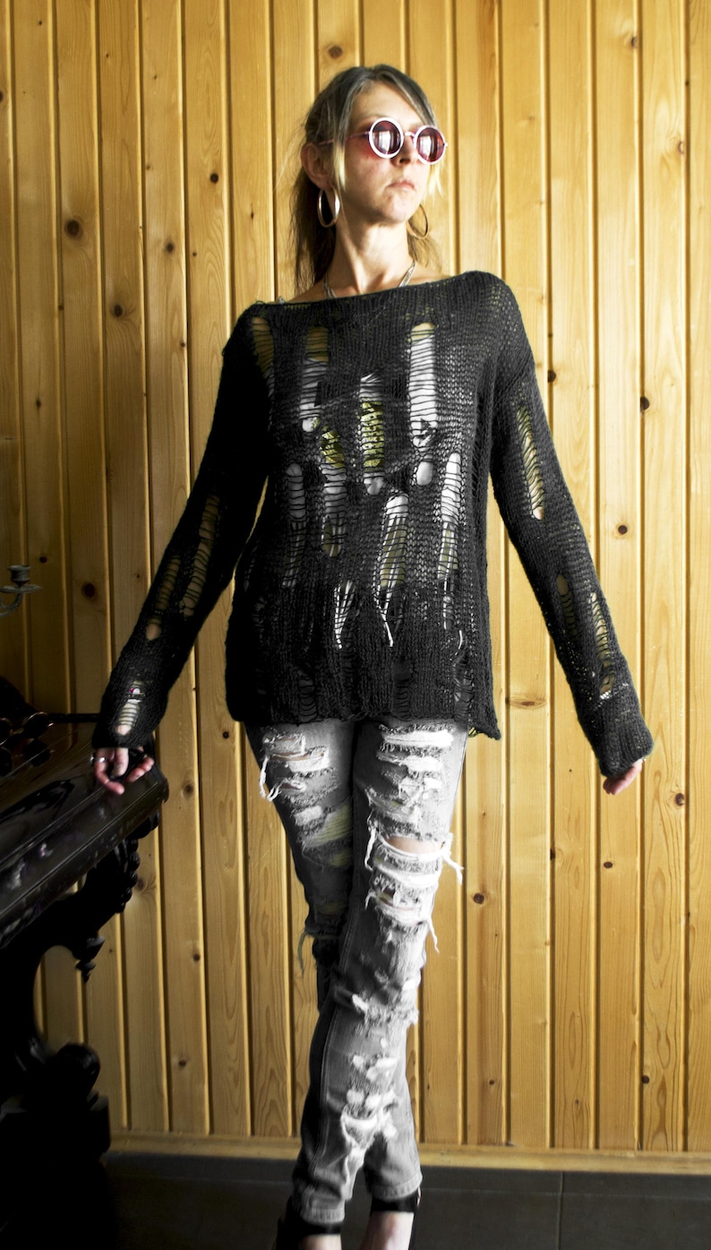 Black open-weave, heavily distressed knit sweater paired with ripped gray jeans; model with pink sunglasses and hoop earrings, wooden wall backdrop