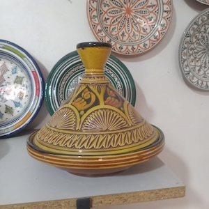 May include: A conical ceramic tagine with a yellow and brown glaze, featuring hand-painted floral and geometric patterns. The lid has a flared top handle and a wide base, decorated with repeating leaf-like motifs and horizontal bands in earthy tones.