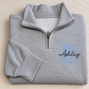 May include: A heather gray quarter-zip sweatshirt featuring a silver metal zipper at the collar and ribbed cuffs. Embroidered on the front in light blue block letters are the initials RN, overlaid with the name Ashley in black cursive script.