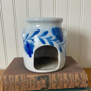 May include: A white ceramic oil warmer featuring a hand-painted blue floral pattern with a wide arched opening at the base. The cylindrical vessel has a smooth glazed finish and is designed with a recessed top section for holding wax or oil.