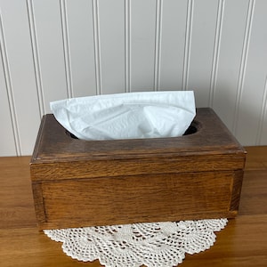 May include: A rectangular dark brown wooden tissue box holder with a visible wood grain texture. The box features a central oval opening on the top surface, from which a single white paper tissue is partially pulled out and displayed.