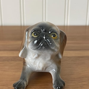 May include: A ceramic figurine of a pug dog with a glossy finish. The dog has a tan and brown coat with a black muzzle and ears. It features prominent yellow eyes, a wrinkled forehead, and is depicted in a seated position with front paws forward.