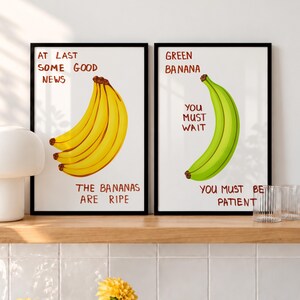 Op de afbeelding: Twee ingelijste wandposters met zwarte lijsten. De linker poster toont gele bananen met de tekst: AT LAST SOME GOOD NEWS THE BANANAS ARE RIPE. De rechter poster toont een groene banaan met de tekst: GREEN BANANA YOU MUST WAIT YOU MUST BE PATIENT.