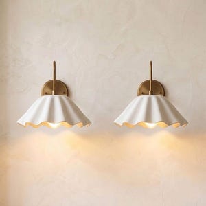 Op de afbeelding: Een paar wandlampen met messingkleurige metalen armen en ronde achterplaten. Elke lamp heeft een brede, witte, gegolfde keramische kap die het licht naar beneden richt en een warme gloed op de muur werpt.