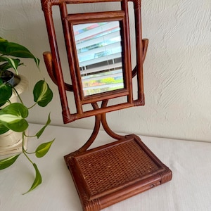 May include: A brown bamboo vanity mirror featuring a rectangular swivel glass pane held within a sturdy frame. The base consists of a flat, woven rattan tray platform supported by curved bamboo legs, providing a stable structure for the mirror.