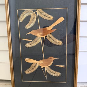 May include: A framed wall art piece featuring two birds crafted from light brown straw or reed inlay against a solid black background. The birds are perched on thin lines with stylized foliage, all enclosed within a simple gold-toned rectangular frame.
