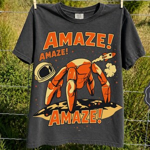 May include: A charcoal gray short-sleeve t-shirt featuring a graphic of an orange robotic crab on a moon surface with an astronaut helmet and a small rocket. The text reads AMAZE! in orange letters above and below the central illustration.