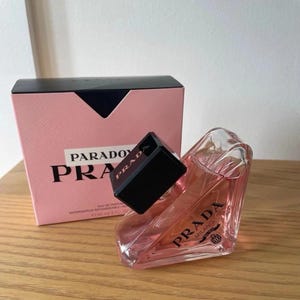 May include: A triangular glass perfume bottle filled with pink liquid, featuring a black cap and the Prada Milano logo. Beside it sits a square pink and black box labeled Paradoxe Prada, with text indicating a volume of 3.0 fl. oz.