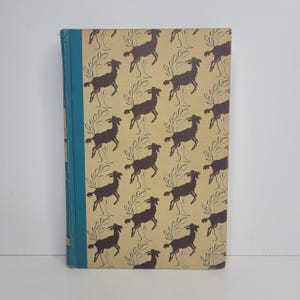 May include: A vintage hardcover book with a tan cover featuring a repeating pattern of brown deer and stylized trees. The spine is a teal blue color. The book is in good condition.