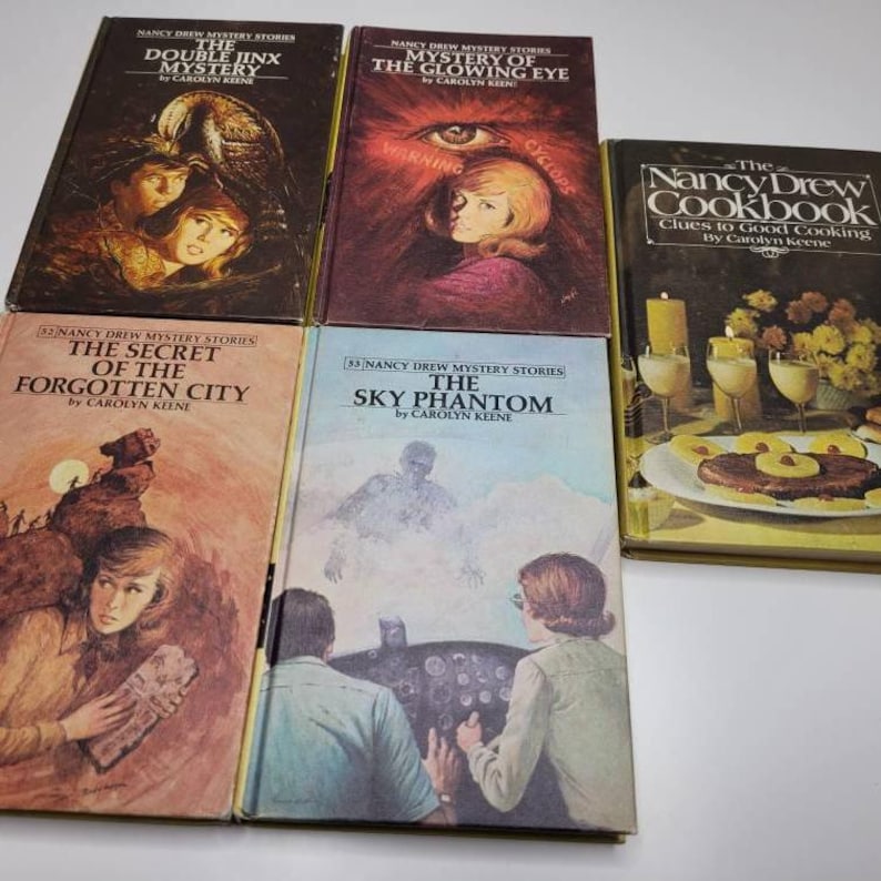 May include: A collection of vintage Nancy Drew mystery books and a cookbook. The book covers feature illustrations with titles like "The Double Jinx Mystery," "The Glowing Eye," and "The Sky Phantom." The cookbook has a yellow cover.