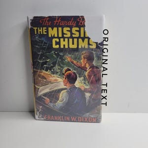 Peut inclure: Livre ancien, "The Hardy Boys: The Missing Chums", avec une illustration de deux jeunes hommes dans une voiture. Le titre est en jaune. Le nom de l'auteur, Franklin W. Dixon, est également visible sur la couverture.