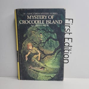 May include: A vintage hardcover book titled "Mystery of Crocodile Island" by Carolyn Keene. The cover features an illustration of a crocodile with a person in a jungle setting. The book has a yellow spine and the words "First Edition" are printed vertically on the right side.