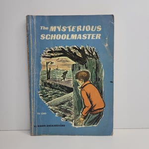 May include: A vintage paperback book titled "The Mysterious Schoolmaster" with a blue cover and an illustration of a boy peeking around a wooden post. The illustration shows a dock, a boat, and a man walking away in the distance.