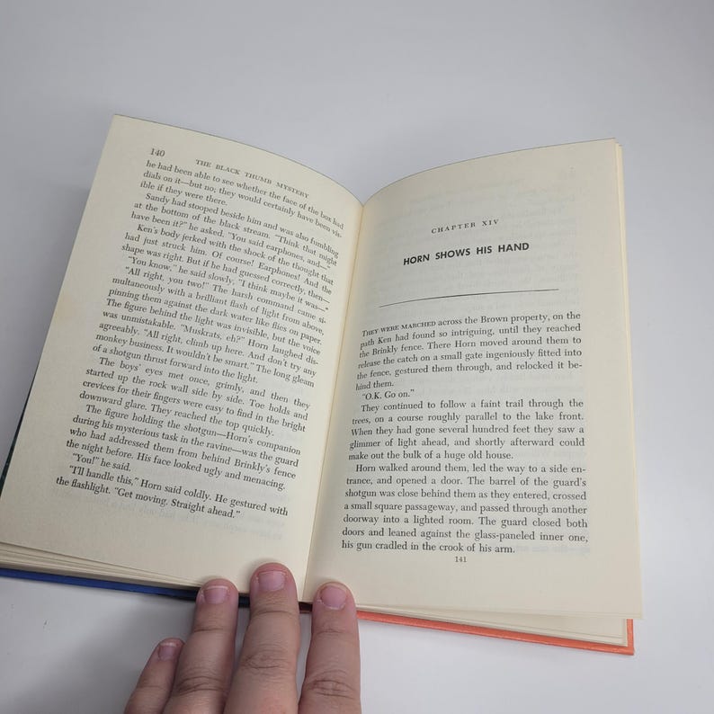 May include: Open book with text on cream-colored pages. The left page has the number 140 at the top, and the right page has the title "HORN SHOWS HIS HAND." A hand is holding the book open, with the text visible.