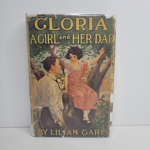 May include: A vintage hardcover book titled Gloria A Girl and Her Dad by Lilian Garis. The cover features a color illustration of a person in a light shirt and dark trousers standing near a tree where a person in a pink dress is sitting.