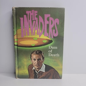 Puede incluir: Una portada de libro de bolsillo con un fondo verde y un platillo volante. El título es "The Invaders" en letras rosas. El libro se titula "Dam of Death" y es una "Edición Autorizada". La portada presenta a un hombre con cabello castaño y una chaqueta marrón.