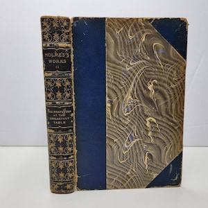 May include: A hardcover book titled Holmes's Works II: The Professor at the Breakfast Table. The spine is black with gold star patterns and lettering. The cover features a dark blue leather-like spine edge and a marbled paper front panel.