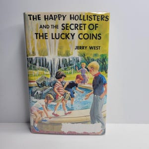 May include: A vintage hardcover book titled "The Happy Hollisters and the Secret of the Lucky Coins" by Jerry West. The cover features an illustration of children playing near a fountain.