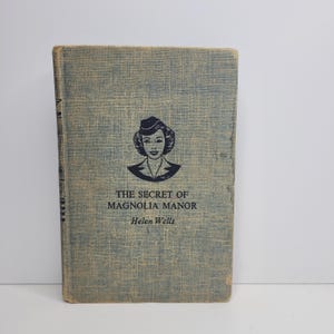 May include: A vintage paperback book with a blue cover and a black and white illustration of a woman wearing a hat. The title of the book is "The Secret of Magnolia Manor" by Helen Wells.
