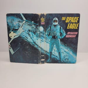 May include: A vintage paperback book cover featuring a blue and black illustration of a spaceman in a silver spacesuit standing in front of a blue and black galaxy background. The book title is "The Space Eagle Operation Doomsday" in white text with a red background.