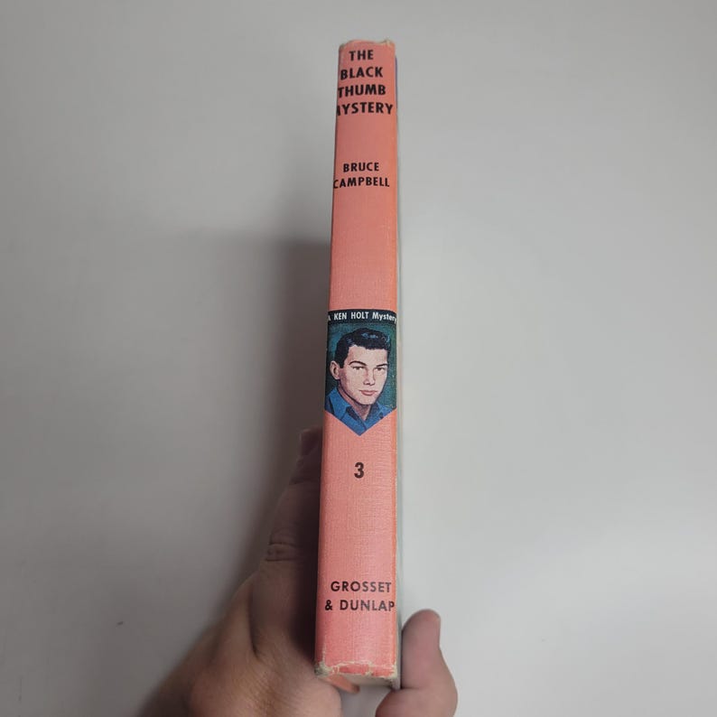 May include: A vintage book titled "The Black Thumb Mystery" by Bruce Campbell. The book has a salmon-colored spine with the author's name and book title in black text. A portrait of a young man is featured on the spine, along with the number 3.
