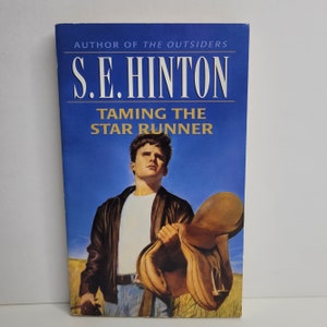May include: A blue paperback book with the title "Taming the Star Runner" by S.E. Hinton. The cover features a young man in a leather jacket holding a saddle.
