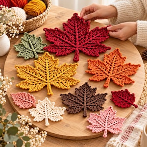 May include: A collection of crocheted maple leaf motifs in various sizes and colors, including deep red, golden yellow, burnt orange, dark brown, cream, and soft pink, arranged on a round wooden surface for decorative display.