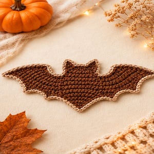 May include: A brown crochet bat applique featuring a dark brown central body and wings with a light beige stitched border. The bat is shaped with pointed ears and scalloped wing edges, showcasing a detailed yarn texture throughout the piece.
