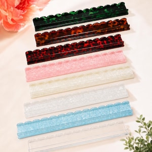 May include: A collection of eight rectangular display stands with scalloped top edges, arranged in a row. The stands feature various finishes including dark green, brown, and red tortoiseshell patterns, solid pink, white, and light blue, and clear.