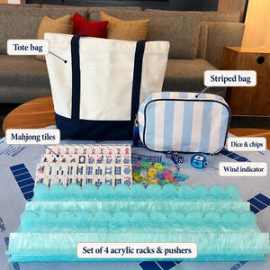 May include: A mahjong set featuring a white and navy blue canvas tote bag, a light blue and white striped zippered bag, a set of four light blue acrylic racks with pushers, a collection of mahjong tiles, colorful plastic chips, dice, and a wind indicator.