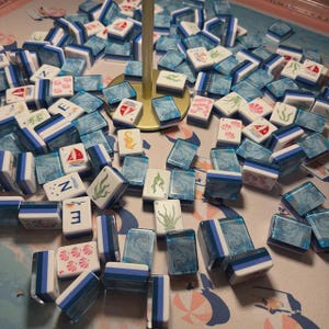 May include: A collection of rectangular mahjong tiles featuring a white face with various nautical and marine-themed illustrations, including sailboats, seashells, and seaweed, paired with a translucent blue marbled back and a gold-colored stand.