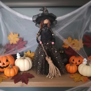 May include: A handcrafted witch doll wearing a black dress with gold star patterns, a matching wide-brimmed lace hat, and a black rose accent at the neckline. The doll holds a small broom made of a wooden twig and natural fibre bristles.