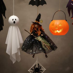 May include: A hanging Halloween decoration set featuring a white ghost with a round head, a witch doll with orange curly hair and a black star-patterned dress holding a broom, an orange plastic jack-o'-lantern, a black bat, and a spider on a web.