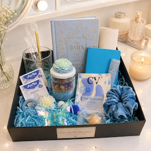 Könnte beinhalten: Eine schwarze Geschenkbox gefüllt mit blauem Papier, die ein Jane Austen Buch, ein Glas mit blauem Blumendeckel, zwei Yogi Tea Bedtime Beutel, ein blaues Notizbuch, CV Augenpads, ein blaues Samt-Haargummi und eine Schmetterlingsspange enthält.