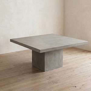 May include: A square, gray concrete table with a solid pedestal base. The table has a minimalist design and is set on a wooden floor. The table's dimensions are approximately 48 inches square.