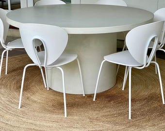 May include: A white round table with a white pedestal base and six white chairs with a unique curved back design. The table and chairs are on a natural fiber rug.