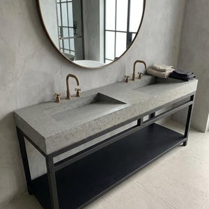 May include: A modern bathroom vanity with a concrete countertop and two sinks. The vanity has a black metal frame and a lower shelf. Gold faucets and a round mirror complete the design. Towels are neatly stacked on the countertop.