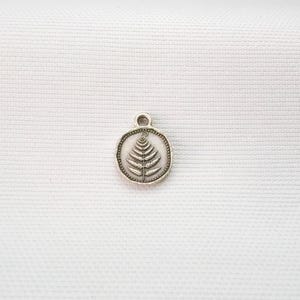 May include: A small, circular silver-toned metal charm featuring an embossed fern leaf design in the center. The charm has a raised border with a beaded texture and a small loop at the top for attachment, presenting a metallic finish.