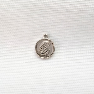 May include: A small, circular silver-toned metal charm featuring an embossed fern leaf design in the center. The charm has a raised beaded border around the edge and a small loop at the top for attachment, showcasing a detailed botanical motif.